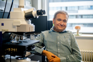 Professor Onur Güntürkün, ein weißer Mann mittleren Alters mit kurzen Haaren, sitzt an einem Schreibtisch mit mehreren Monitoren und einem Mikroskop. © RUB/Marquard
