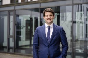 Ein Mann in Anzug und Krawatte (Tobias Ruck) steht selbstbewusst vor einem modernen Gebäude. © RUB/Marquard