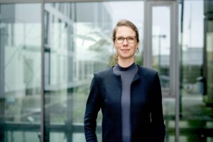 Eine Frau mit Brille steht vor der Kulisse eines reflektierenden Glasgebäudes. © RUB/Marquard