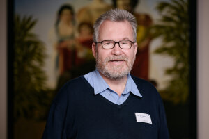 Bild von Prof. Juckel, der eine Brille und einen Pullover trägt, steht vor einem Gemälde.