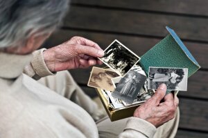 Eine ältere Frau mit grauem Haar hält eine Schachtel mit historischen Fotografien, die ein Brautpaar, ein Familienporträt bei einer Hochzeit, ein Einzelporträt sowie eine Mutter mit ihrem Kind zeigen.