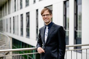 Ein Mann, Caspar Schwiedrzik, in einem Anzug und mit Brille steht vor einem modernen Gebäude. © Michael Schwettmann