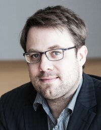Profilbild von Prof. Dr. Jonas Rose, ein Mann mit Dreitagebart, Brille und dunkelblonden kurzen Haaren, der ein gestreiftes Hemd und ein dunkles Jackett trägt.
